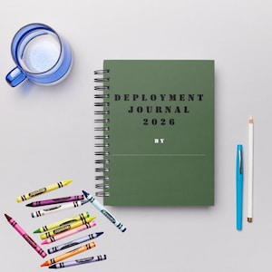 Peut inclure: Un journal vert à spirales avec le texte "DEPLOYMENT JOURNAL 2026" et une collection de crayons de couleur. Un stylo bleu et un crayon blanc sont également présents. Un verre bleu et blanc est dans le coin supérieur gauche.