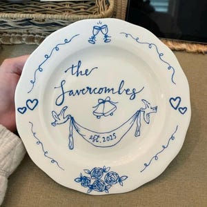 May include: White ceramic plate with a scalloped edge, featuring blue hand-painted details. The plate is inscribed with "the Savercombes" and "est. 2025", along with decorative elements like hearts, doves, and floral designs. A charming wedding or anniversary keepsake.