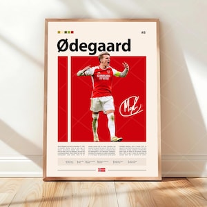 May include: A framed poster featuring a photograph of soccer player Martin Ødegaard in a red and white jersey against a red background. The text includes his name, the number 8, a signature, and biographical details in black on a cream background.