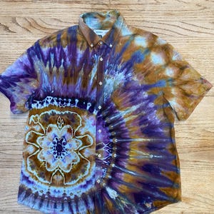 May include: A short-sleeve button-down shirt featuring a tie-dye pattern in shades of purple, brown, and white. The design includes a prominent circular mandala-like motif on the lower left side, a button-down collar, and a single chest pocket.
