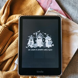 May include: Kindle e-reader with a clear protective case. The screen displays a graphic of three cartoon ghosts with the text "SIP, READ & SPOOKY VIBES ONLY." The e-reader is on a textured surface with a gold sweater and a marble-patterned book cover.