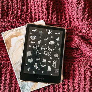 Pode incluir: Um leitor de e-books Kindle com uma capa transparente exibindo o texto "All booked for Fall" e ícones de outono. Descansa sobre um caderno com padrão de mármore e um cobertor de malha bordô.