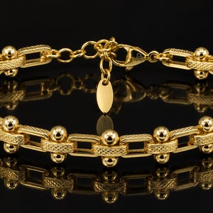 May include: A gold-toned chain bracelet featuring alternating rectangular links with a cross-hatched texture and smooth, polished spherical beads. The bracelet includes a lobster claw clasp, a short extender chain, and a small oval charm.