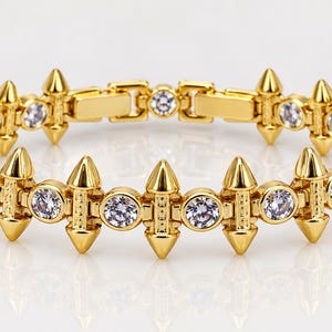 May include: A gold-tone link bracelet featuring alternating circular clear stones and pointed, cone-shaped metal spikes. The bracelet is constructed with a series of connected segments, each holding a round stone and a vertical spike element.