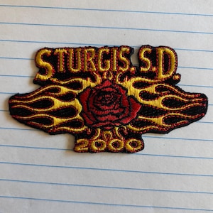 Puede incluir: Parche bordado con el texto "STURGIS S.D." en dorado, una rosa roja y el año "2000". El diseño incluye llamas negras y doradas. Adecuado para chaquetas o bolsos.