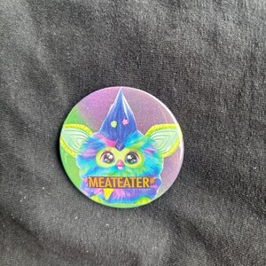 Puede incluir: Un botón redondo con un diseño de criatura de dibujos animados colorida. La criatura tiene pelo azul, verde y morado, ojos grandes y un sombrero puntiagudo. El botón incluye la palabra "MEATEATER" en una pancarta amarilla, sobre un fondo morado y verde.