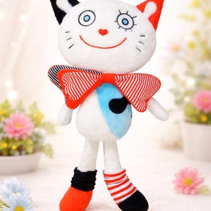 May include: A soft toy cat with a white body, black and red ears, and a red bow tie. The doll has a smiling face with blue eyes and a red heart nose. It wears black and red striped socks and orange shoes.