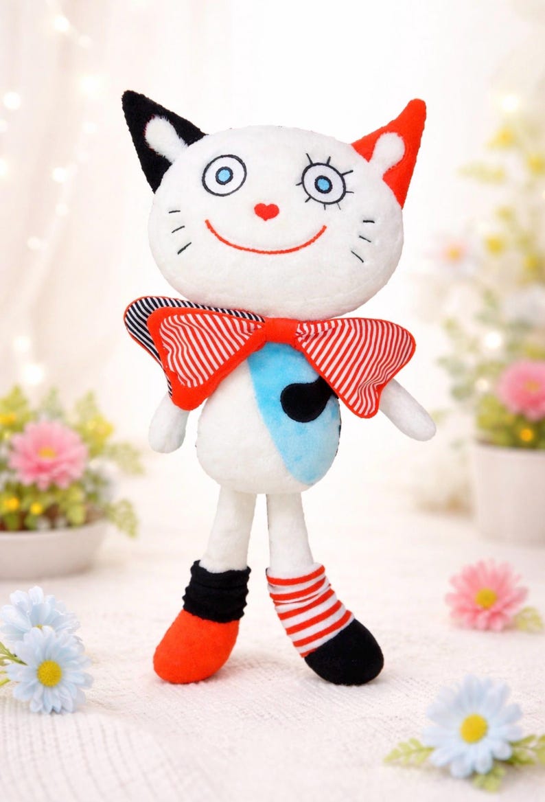 May include: A soft toy cat with a white body, black and red ears, and a red bow tie. The doll has a smiling face with blue eyes and a red heart nose. It wears black and red striped socks and orange shoes.