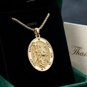 May include: A gold-toned oval pendant necklace featuring an embossed image of Saint Raphael and the text "Saint Raphael Pray For Us". The pendant is displayed inside a dark green velvet jewellery box next to a small card with "Thank You" printed on it.