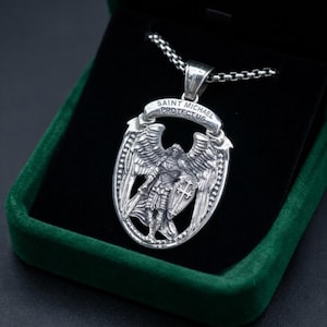 May include: A silver-toned metal pendant featuring an embossed figure of Saint Michael with wings, holding a shield and sword. The pendant is oval-shaped with a banner at the top inscribed with the text SAINT MICHAEL PROTECTUS and hangs on a chain.