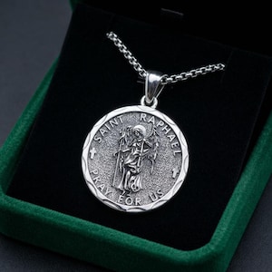 May include: A silver-toned circular pendant necklace featuring an embossed figure of Saint Raphael with wings. The text SAINT RAPHAEL PRAY FOR US is engraved around the perimeter. The pendant hangs from a silver-toned box chain link necklace.