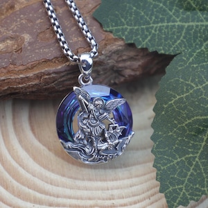 May include: A silver pendant necklace with a round, blue crystal and an angel figure. The angel has wings and holds a scale. The chain is a silver box chain.