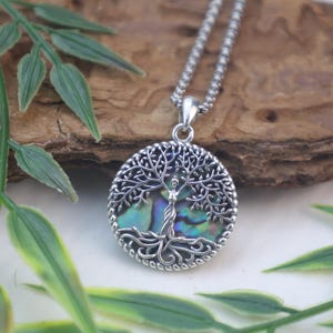 Puede incluir: Collar con colgante plateado con un diseño detallado del árbol de la vida. El colgante tiene un marco circular con un borde texturizado, y las ramas y raíces del árbol están intrincadamente elaboradas. El centro tiene una incrustación de concha de abulón iridiscente.