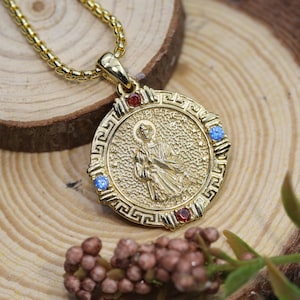 Puede incluir: Un collar con colgante dorado con un medallón circular que representa una figura religiosa. El medallón está adornado con piedras preciosas rojas y azules y un borde de clave griega. La cadena también es dorada.