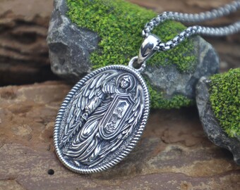 Può includere: Collana con pendente ovale argentato con un rilievo dettagliato di un angelo che tiene uno scudo. Il pendente è sospeso a una catena abbinata. Il design presenta dettagli intricati e un bordo strutturato, adatto per uso religioso o simbolico.