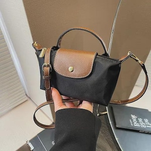 May include: Black handbag with a brown flap and handle. The bag has a gold zipper and buckle, and a brown adjustable shoulder strap. The bag is held by a person.