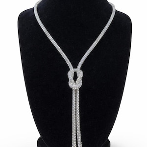 May include: A silver lariat necklace on a black mannequin. The necklace has a knotted centre and two long, dangling strands. It's made of small, reflective beads, creating a sparkling effect. The necklace is a stylish accessory.