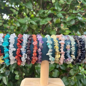 May include: A display of gemstone chip bracelets in various colors, including turquoise, red, white, and purple. The bracelets are arranged on a teal velvet-covered cylinder with gold accents, set against a backdrop of green foliage.