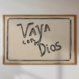 May include: A rectangular framed wall art piece featuring a light beige background with a black hand-drawn border. The text Vaya con Dios is written in the centre in a black, sketchy, handwritten font style, enclosed within the rectangular frame.