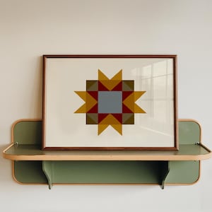 Puede incluir: Una lámina enmarcada con un patrón geométrico de estrella en tonos amarillo mostaza, rojo oscuro, verde oliva y un cuadrado central gris. La obra se exhibe sobre un estante de pared verde salvia con bordes redondeados y moldura de madera clara.