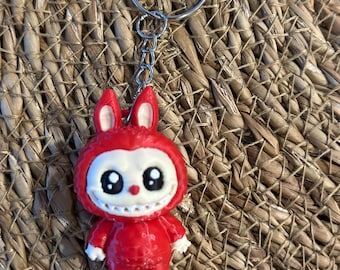 May include: A red keychain featuring a cartoonish figure with a white face, large black eyes, and a toothy grin. The figure has small white hands and feet. The keychain includes a silver metal ring and chain. The background is a woven, textured surface.