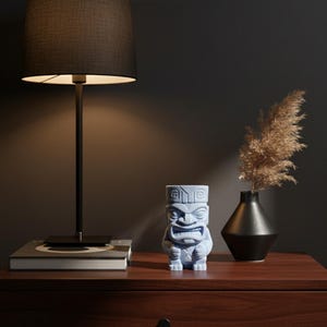 May include: A blue tiki statue with a detailed carved design sits on a wooden surface next to a black lamp and a black vase with dried plants. The lamp has a black lampshade and a metal stand.