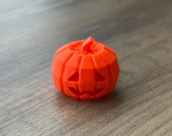 Puede incluir: Una figura de calabaza de color naranja brillante impresa en 3D, de estilo low-poly, que presenta una cara de linterna tallada con ojos triangulares y una boca dentada. El objeto tiene una textura de superficie geométrica facetada y un pequeño tallo.