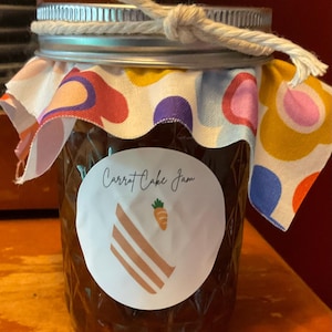 May include: A jar of carrot cake jam with a white label featuring the text "Carrot Cake Jam" and a slice of cake graphic with a carrot. The jar is sealed with a metal lid, a patterned fabric, and tied with a twine bow.