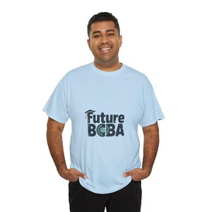 Può includere: T-shirt azzurra con la scritta "Future BCBA" in blu navy, con un cappello di laurea e un design a corona d'alloro. La maglietta è realizzata in un materiale morbido.