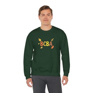 Puede incluir: Una sudadera verde oscuro con las letras "BCBA" en naranja, marrón y amarillo, rodeadas de gráficos de hojas. La sudadera es de cuello redondo.