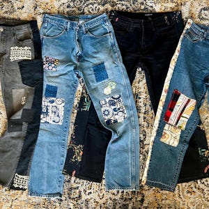 May include: A collection of five pairs of jeans with unique patchwork designs. The jeans vary in color, including light and dark washes, and feature decorative patches in various patterns and textures. Some have lace or fabric accents.
