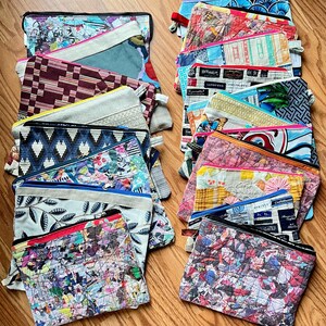 May include: A collection of rectangular zipped pouches featuring a variety of colourful fabric patterns, including floral, geometric, and abstract prints. Each pouch has a top zip closure and is constructed from quilted or layered textile materials.