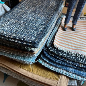 May include: A stack of fabric samples in various colors and textures. The samples include blue, beige, and patterned fabrics. One sample is a small rug with orange and white stripes. The fabrics are stacked on top of each other.