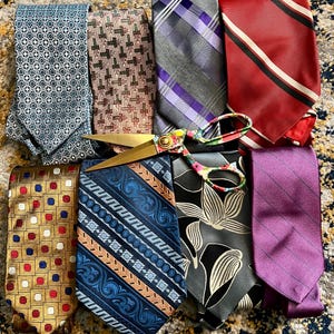 May include: A collection of men's ties in various patterns and colors, including blue, red, purple, and gold. A pair of gold scissors with floral handles rests on top of the ties. The ties have geometric, striped, and floral designs.
