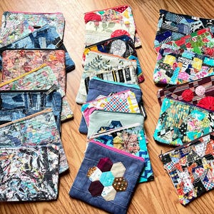 Könnte beinhalten: Eine Sammlung rechteckiger Stofftaschen mit verschiedenen Patchwork-Designs, gesteppten Texturen und farbenfrohen Mustern. Jede Tasche verfügt über einen Reißverschluss und zeigt einzigartige Kombinationen aus floralen, geometrischen und abstrakten Drucken.