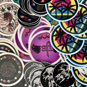May include: A collection of embroidered patches featuring various designs, including a bird, palm trees, and a skull. The patches are circular and have a white background with colorful embroidery. The patches are arranged in a pile on a table.