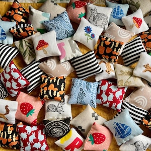 May include: A collection of small, square fabric sachets featuring diverse patterns including mushrooms, strawberries, rabbits, ships, squirrels, rainbows, and botanical prints in various colors like black, white, red, blue, and orange.