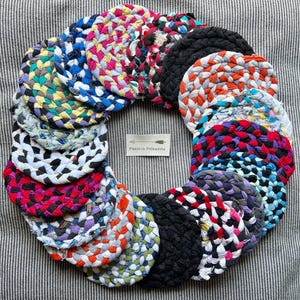 May include: A collection of colorful, round, braided fabric coasters arranged in a circular shape. The coasters feature various colors, including blue, red, black, and white. The image includes a small tag with the text "Panie in Polkadots".