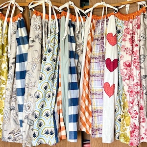 May include: A collection of hanging fabric aprons featuring various patterns including yellow floral, blue and white gingham, geometric fan shapes, orange plaid, and a white apron with three red heart appliques and pink floral prints.