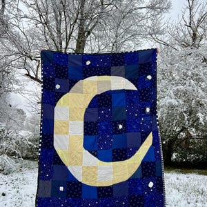 May include: A handmade quilt with a large crescent moon design in yellow and white fabric. The quilt is made of patchwork squares in shades of blue and features white stars scattered across the background.