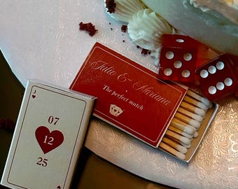 Può includere: Una composizione decorativa con una torta bianca glassata e briciole di velluto rosso. Una scatola di fiammiferi rossa, con la scritta "Julie & Mariana" e "The perfect match", è aperta, rivelando i fiammiferi. Sono presenti anche una carta da gioco e dadi rossi.