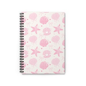Pode incluir: Um caderno espiral com fundo branco e um padrão repetido de conchas rosa, estrelas do mar, laços e corações. A capa do caderno apresenta um design feminino e caprichoso, ideal para anotações ou diários.