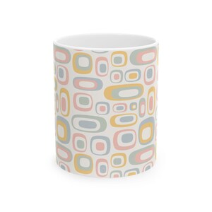 May include: White ceramic mug with a repeating pattern of retro-style shapes in pastel colors: pink, yellow, gray, and blue. The design features rounded rectangles and ovals, creating a mid-century modern aesthetic.