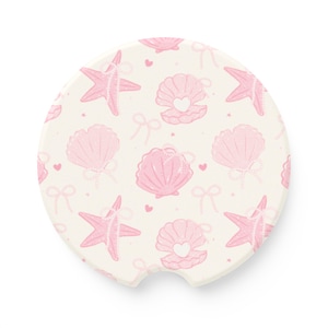 May include: Round car coaster with a cream background and a repeating pattern of pink seashells, starfish, bows, and hearts. The seashells have a pearl strand and a heart in the center. The starfish also have pearl strands.
