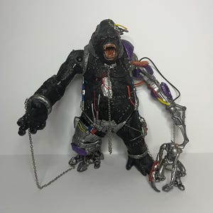 May include: A black gorilla action figure with cybernetic enhancements. The figure has an open mouth, showing teeth, and is holding a chain. The figure has silver, purple, and yellow accents.