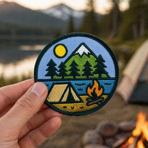 May include: A circular embroidered patch featuring a light blue sky with a yellow sun, a green mountain with a white peak, dark green pine trees, a blue lake, a tan tent, and an orange campfire on a tan ground, all enclosed by a dark border.