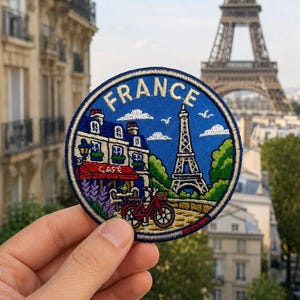May include: A circular embroidered patch featuring a blue background with the word FRANCE in gold letters. The design depicts a Parisian cafe with a red awning, a red bicycle, green trees, and the Eiffel Tower under a blue sky with white clouds.