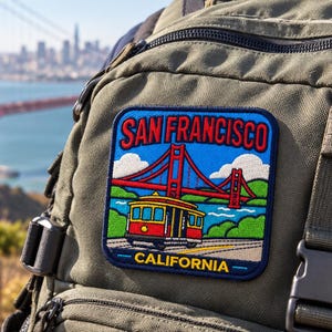 May include: An olive green backpack featuring a rectangular embroidered patch. The patch depicts a red cable car, the Golden Gate Bridge, and blue sky with white clouds, with the text SAN FRANCISCO and CALIFORNIA in yellow lettering.
