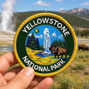 May include: A circular embroidered patch featuring a dark green border with a yellow edge. The center depicts a landscape with a geyser, a brown bison, green pine trees, and blue mountains under a yellow sun. The text reads YELLOWSTONE NATIONAL PARK.