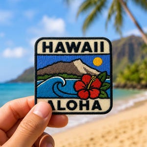 May include: A square embroidered patch featuring the text HAWAII at the top and ALOHA at the bottom. The design depicts a brown mountain, a blue ocean wave, a yellow sun, and a red hibiscus flower with green leaves against a blue sky background.